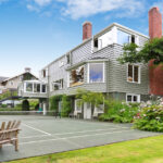 A modernly built house with a tennis court in the backyard. The backyard is beautifully landscaped with trees and plants.