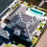 A technician is working on solar panels installed on a luxury waterfront home under a clear, sunny sky.