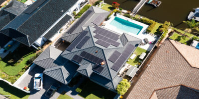 A technician is working on solar panels installed on a luxury waterfront home under a clear, sunny sky.