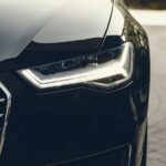 Tips To Care for Your Audi Vehicle Correctly A close-up of a headlight on a black vehicle parked on a gray brick pathway. The background is blurry.