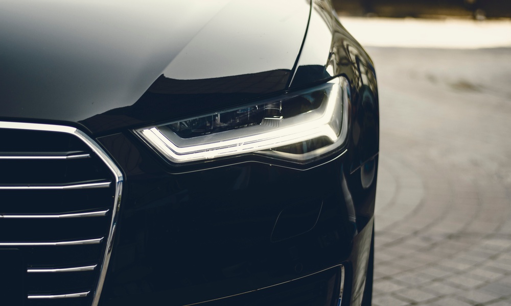 A close-up of a headlight on a black vehicle parked on a gray brick pathway. The background is blurry.