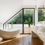 Signs It’s Time To Give Yourself a More Luxurious Bathroom A clean, airy bathroom with a freestanding bathtub and a floor-to-ceiling window leading to a deck or patio.