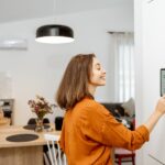 Things You Need Before Starting Your Smart Home A smiling woman in an orange blouse presses the screen of a large touchscreen that hangs on the wall of a home.