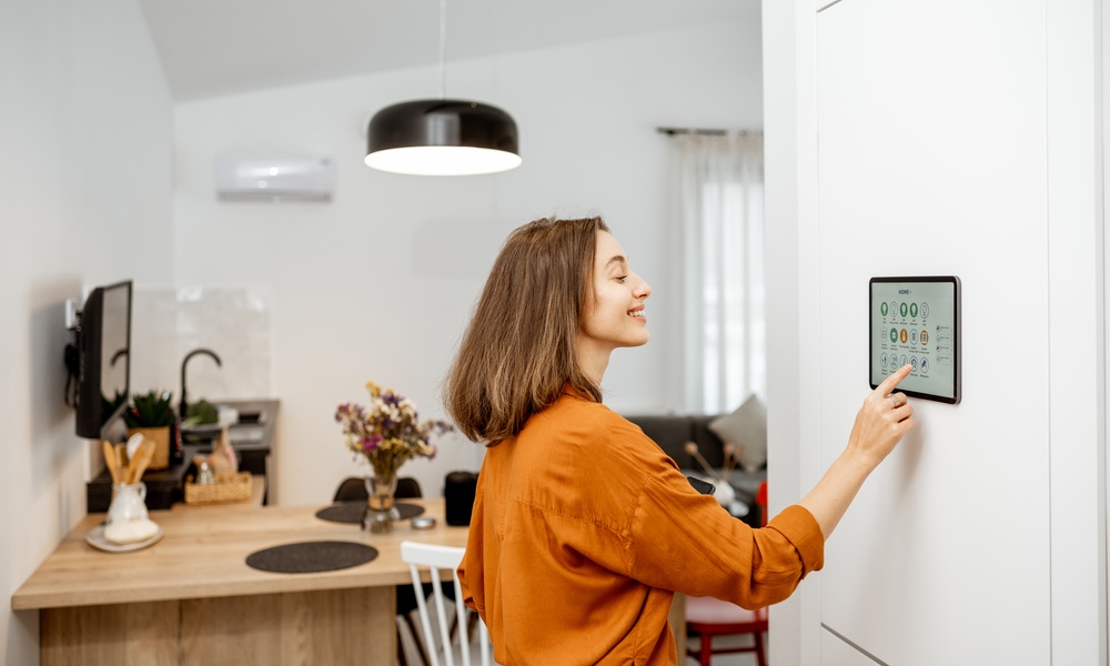 A smiling woman in an orange blouse presses the screen of a large touchscreen that hangs on the wall of a home.