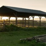 5 Interesting Things You Can Do With a Pole Barn An old pole barn is in front of a fence and a pile of logs with the sun and mountains in the background.