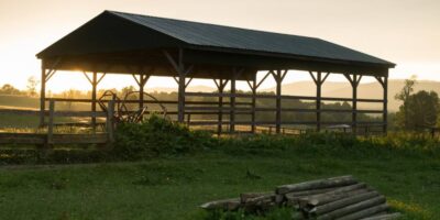 An old pole barn is in front of a fence and a pile of logs with the sun and mountains in the background.