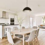 A dining table with six gray chairs stands in front of a kitchen with white cabinets and black countertops.