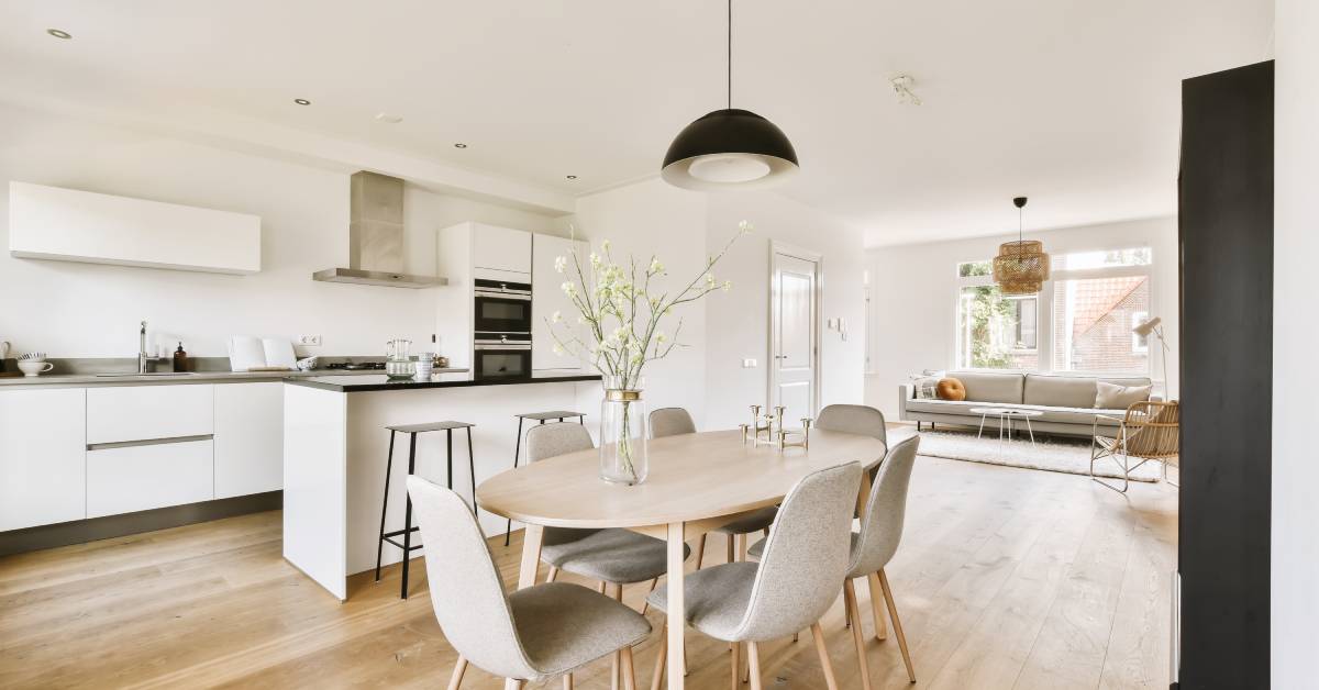 A dining table with six gray chairs stands in front of a kitchen with white cabinets and black countertops.