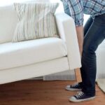 A man wearing a blue plaid shirt, jeans, and sneakers carrying an off-white sofa across a hardwood floor.