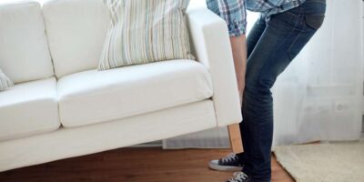 A man wearing a blue plaid shirt, jeans, and sneakers carrying an off-white sofa across a hardwood floor.