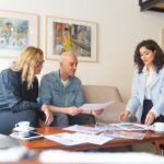 Two women and a man sit on a couch and chairs, looking at papers that sit on a wooden coffee table.