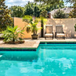 Closeup of a luxurious backyard pool with bright turquoise water next to a concrete patio with chairs and plants.