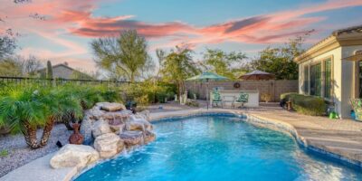 A circular backyard pool with a stone water feature surrounded by landscaping and two chairs and umbrellas.