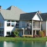 Lake Home Tips: How To Maintain Your Home in the Off-Season A beautiful luxury home sits on the edge of a lakefront property with gorgeous landscaping and blue skies in the background.