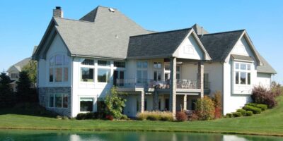 A beautiful luxury home sits on the edge of a lakefront property with gorgeous landscaping and blue skies in the background.