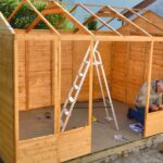 Natural Constructions: Saving Money on Your Wood Building An older man wearing a jacket and jeans kneeling down with a power tool while working on a wooden building.