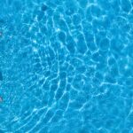 A woman sitting in a pink flotation ring in a pool wearing a multicolor swimsuit with vertical stripes and a sun hat.