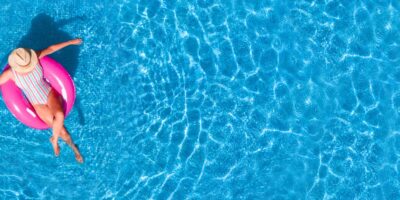 A woman sitting in a pink flotation ring in a pool wearing a multicolor swimsuit with vertical stripes and a sun hat.