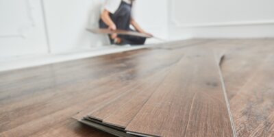 A stack of luxury vinyl flooring planks is in focus in the foreground, and a worker is installing the floor in the background.