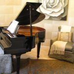 A grand piano sits in a rounded part of a room with other furniture and accessories, including a rose painting behind it.