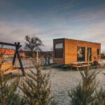 Debunking 4 Common Misconceptions About Tiny Homes A tiny wooden home with large windows parked on a camping site with a picnic table and swinging bench next to it.