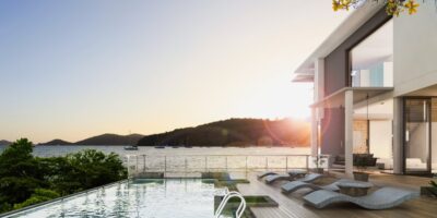 The sun sets on a modern, luxury beach house with a large rectangular pool. Sailboats float on the ocean in the background.