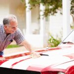 An older man is washing his vintage red luxury sports car with white stripes. He's applying wax to the hood.