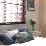 6 Tips for Setting Up Your Guest Bedroom A bedroom with a large window, a made bed with teal bedding, a side table with flowers, artwork, and a floor lamp.