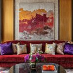 5 Art Lighting Tips To Elevate Your Home Display A red velvet sofa with purple and patterned pillows sits below abstract art and gold sconces on the wall.