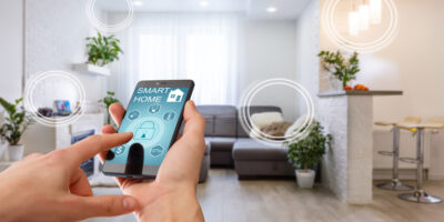 A hand holding a smartphone about to press a lock button in their home's living room. The phone reads "smart home".