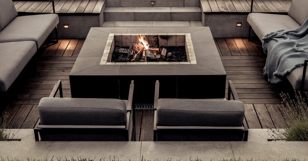 Gray stone steps lead to a sunken patio with a lit fire pit, metal seating, and distressed wooden flooring.