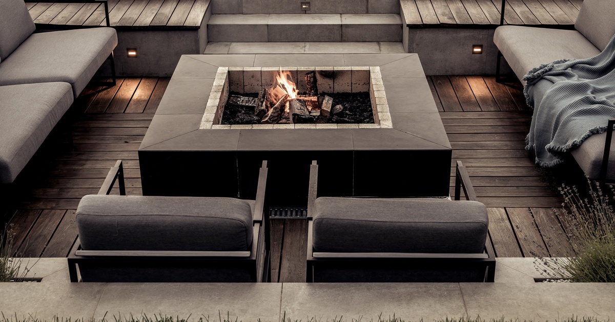Gray stone steps lead to a sunken patio with a lit fire pit, metal seating, and distressed wooden flooring.