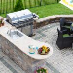 A patio in the backyard of a home with a grill, countertop, and table for dining in front of a black fence.